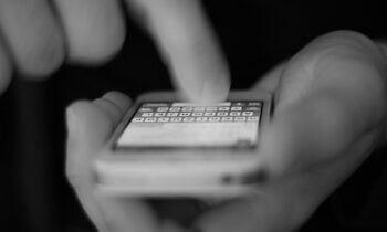 Primo piano in bianco e nero di mani che tengono un cellulare e digitano sulla tastiera, immagine che introduce il tema dello smishing.