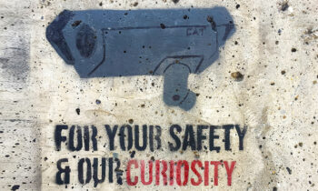 Disegno a spray su un muro di una telecamera di videosorveglianza grigia con sotto la scritta "For your safety and our curiosity", immagine che introduce i pericoli per la privacy legati alle nuove tecnologie di videosorveglianza AI based.