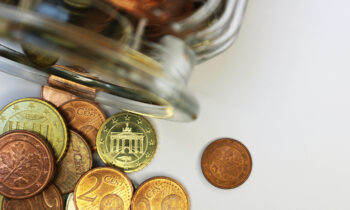 Barattolo di vetro appoggiato su un piano bianco, dal quale escono degli euro in moneta, immagine che introduce il tema dei costi dei sistemi di sicurezza.